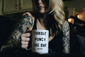 tattooed woman sitting on a couch holding a mug that says throat punch the day