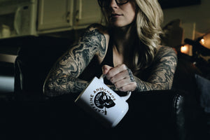 tattooed woman sitting on a couch holding a white mug that says caffeine and chaos