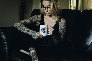 tattooed woman sitting on a couch holding a mug with a skull design
